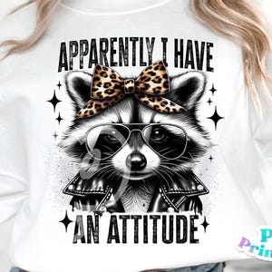 May include: A white sweatshirt featuring a black and white illustration of a raccoon wearing a black leather jacket, sunglasses, and a leopard print bow. The text "APPARENTLY I HAVE AN ATTITUDE" is printed in black on the sweatshirt.