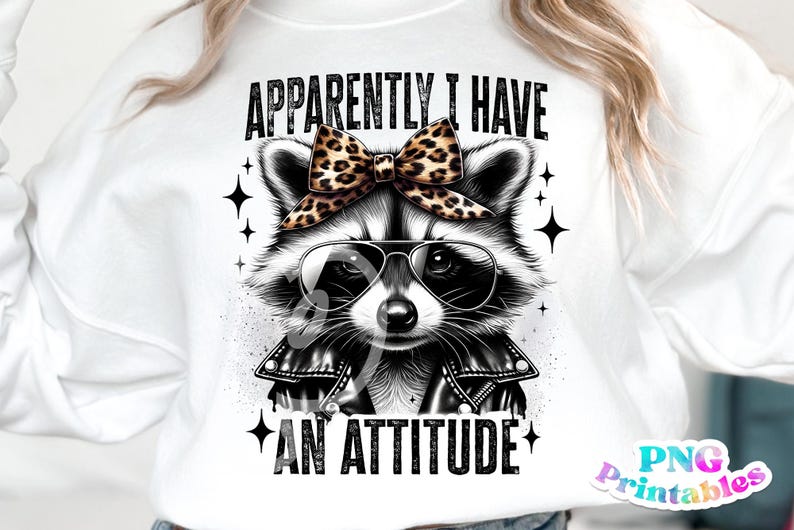 May include: A white sweatshirt featuring a black and white illustration of a raccoon wearing a black leather jacket, sunglasses, and a leopard print bow. The text "APPARENTLY I HAVE AN ATTITUDE" is printed in black on the sweatshirt.