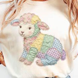 May include: A cream-coloured t-shirt displaying a patchwork lamb design in pastel shades. The lamb has a pink face, ears, and feet, with a floral patterned headpiece. The patchwork includes blue, yellow, and purple squares.
