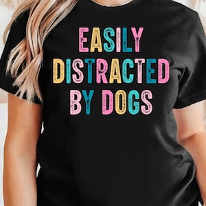 Może przedstawiać: Czarny T-shirt z napisem "Easily Distracted By Dogs" w kolorowych literach.