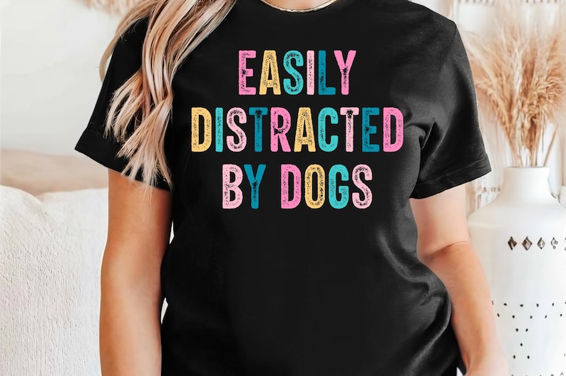 Może przedstawiać: Czarny T-shirt z napisem "Easily Distracted By Dogs" w kolorowych literach.