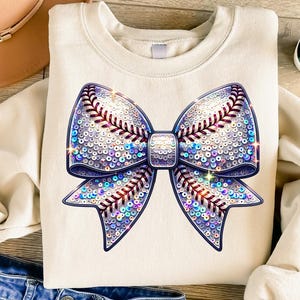 May include: A white sweatshirt featuring a large, sparkly, iridescent bow. The bow is made of sequins and has a baseball stitch design.