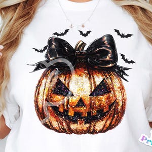 Puede incluir: Una camiseta blanca con una calabaza naranja brillante con un lazo negro y murciélagos impresos en ella.
