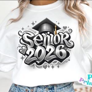 K&ouml;nnte beinhalten: Wei&szlig;es Sweatshirt mit einem schwarz-grauen Grafikdesign. Das Design zeigt den Text "Senior 2026" in einer stilisierten Schriftart, eine Abschlusskappe, Herzen und Sterne. Eine goldene Quaste h&auml;ngt an der Kappe.