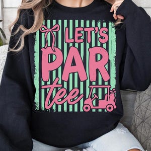 May include: Black sweatshirt featuring the text "Let's Par Tee" in pink, with a golf cart and bow graphic. The design has green and black vertical stripes. A casual top for golf lovers. The measurements are in centimeters.