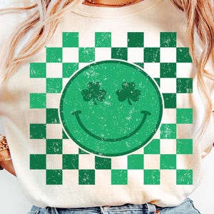 May include: Cream-colored sweatshirt featuring a green smiley face with shamrock eyes and a smiling mouth. The smiley face is set against a green and white checkered background. The design has a distressed, vintage look.