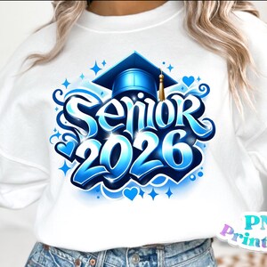 K&ouml;nnte beinhalten: Wei&szlig;es Sweatshirt mit einem blau-wei&szlig;en Aufdruck mit der Aufschrift "Senior 2026". Das Design beinhaltet eine Abschlusskappe, Herzen und Sterne. Das Sweatshirt hat einen Rundhalsausschnitt.