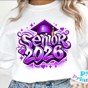 K&ouml;nnte beinhalten: Wei&szlig;es Sweatshirt mit einem lila-wei&szlig;en Aufdruck "Senior 2026". Das Design beinhaltet eine Abschlusskappe, Herzen und Sterne. Der Text ist in einer stilisierten Schriftart mit Farbverlauf gehalten.
