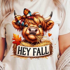 Può includere: Una maglietta bianca con un'illustrazione di una mucca Highland marrone e bianca che indossa un fiocco a quadri. La mucca sta mangiando fieno e c'è un cartello che dice "Hey Fall" davanti a lei.