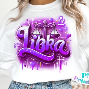 Pode incluir: Uma camisa de moletom branca com um design gr&aacute;fico roxo e dourado que apresenta a palavra "Libka" e um s&iacute;mbolo do zod&iacute;aco Libra. O design inclui cora&ccedil;&otilde;es, estrelas e gotas de tinta.