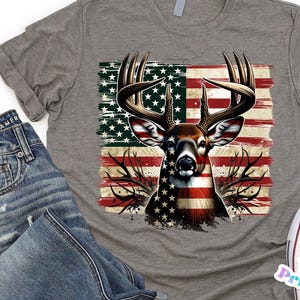 May include: A heather gray t-shirt featuring a deer design with an American flag motif. The deer's body is colored with the stars and stripes. The shirt is paired with blue jeans and white sneakers.