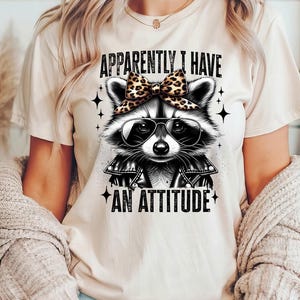 May include: A white t-shirt featuring a black and white illustration of a raccoon wearing sunglasses and a leopard print bow. The text "Apparently I Have An Attitude" is printed below the raccoon.