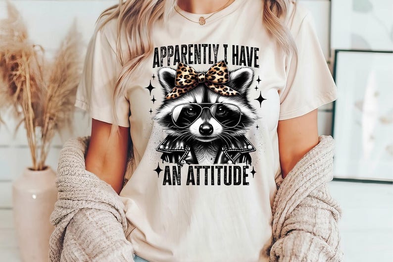 May include: A white t-shirt featuring a black and white illustration of a raccoon wearing sunglasses and a leopard print bow. The text "Apparently I Have An Attitude" is printed below the raccoon.