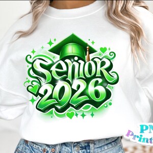 K&ouml;nnte beinhalten: Wei&szlig;es Sweatshirt mit einem leuchtend gr&uuml;nen "Senior 2026" Aufdruck. Das Design beinhaltet eine Abschlusskappe, Herzen und dekorative Wirbel. Der Text ist in einer fetten, stilisierten Schriftart gehalten, ideal f&uuml;r Abschlussfeiern.