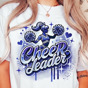 May include: White t-shirt with a blue and purple graphic design of a cheerleader holding pom-poms. The text "Cheerleader" is written in a stylized font.