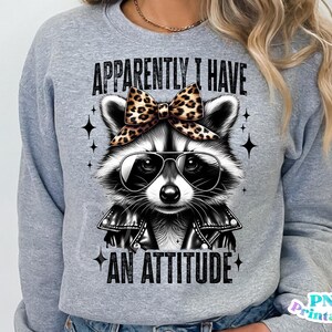 May include: Grey sweatshirt featuring a black and white raccoon wearing a leopard print bow and sunglasses. The text "Apparently I Have An Attitude" is printed on the sweatshirt.