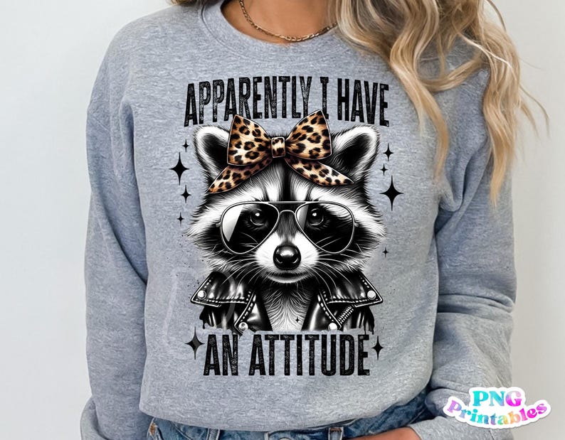 May include: Grey sweatshirt featuring a black and white raccoon wearing a leopard print bow and sunglasses. The text "Apparently I Have An Attitude" is printed on the sweatshirt.