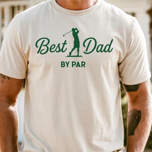 K&ouml;nnte beinhalten: Cremefarbenes T-Shirt mit dem Text "Best Dad" in gr&uuml;ner, kursiver Schrift und einer gr&uuml;nen Silhouette eines Golfspielers. Darunter stehen die Worte "BY PAR" in gr&uuml;nen Blockbuchstaben. Das T-Shirt hat kurze &Auml;rmel.