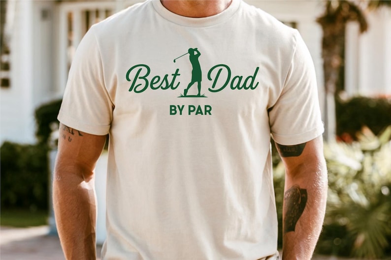 K&ouml;nnte beinhalten: Cremefarbenes T-Shirt mit dem Text "Best Dad" in gr&uuml;ner, kursiver Schrift und einer gr&uuml;nen Silhouette eines Golfspielers. Darunter stehen die Worte "BY PAR" in gr&uuml;nen Blockbuchstaben. Das T-Shirt hat kurze &Auml;rmel.