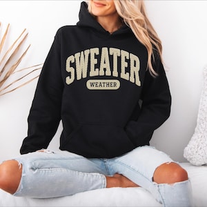 May include: Black hooded sweatshirt with the text "SWEATER WEATHER" in a cream-coloured, collegiate-style font. The sweatshirt has a front pocket and a drawstring hood. The model is wearing light blue jeans.