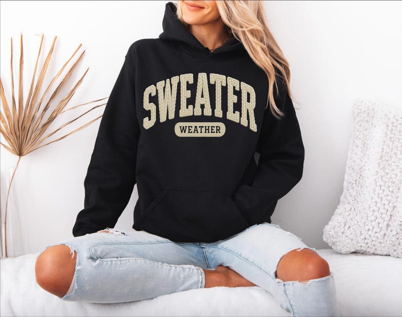 May include: Black hooded sweatshirt with the text "SWEATER WEATHER" in a cream-coloured, collegiate-style font. The sweatshirt has a front pocket and a drawstring hood. The model is wearing light blue jeans.
