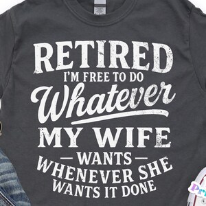 May include: Dark grey t-shirt with the text "RETIRED I'M FREE TO DO WHATEVER MY WIFE WANTS WHENEVER SHE WANTS IT DONE" in white. The shirt is styled with sunglasses, jeans, and white trainers.