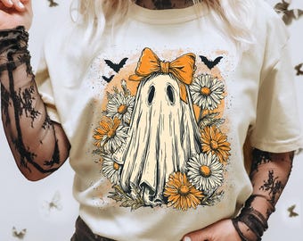 May include: Cream-colored t-shirt featuring a ghost design with an orange bow, surrounded by daisies and bats. The graphic has a vintage, hand-drawn style. The shirt is a casual, everyday wear item.