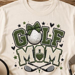 May include: A cream-colored t-shirt featuring a graphic design with the text GOLF MOM in textured green and white varsity-style letters. A white golf ball with a green bow sits above the text, and two crossed golf clubs are positioned below.