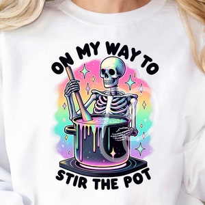 Puede incluir: Una sudadera blanca con un gráfico de arcoíris de un esqueleto revolviendo una olla con el texto "On my way to stir the pot."