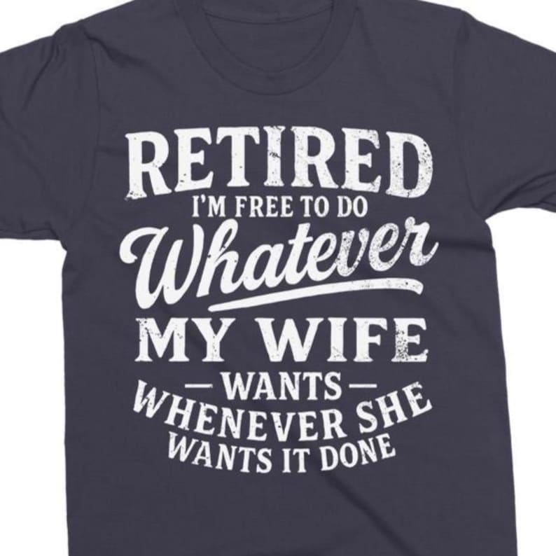 May include: Dark blue t-shirt with white text. The text says "RETIRED I'M FREE TO DO WHATEVER MY WIFE WANTS WHENEVER SHE WANTS IT DONE". The shirt has a crew neck and short sleeves.