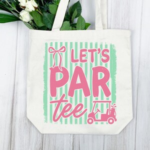 May include: Cream tote bag with the pink text "Let's Par Tee", a golf cart graphic, and a bow. The background has green stripes. White flowers and greenery are visible at the top.