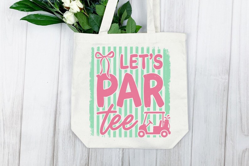 May include: Cream tote bag with the pink text "Let's Par Tee", a golf cart graphic, and a bow. The background has green stripes. White flowers and greenery are visible at the top.