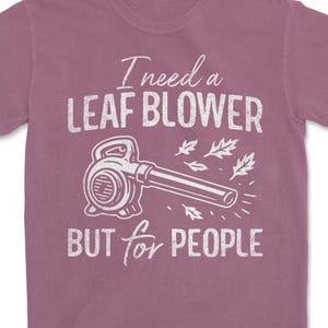 Peut inclure: T-shirt mauve avec un dessin blanc de souffleur de feuilles et le texte "I need a LEAF BLOWER BUT for PEOPLE". Le design comprend des feuilles stylisées, parfait pour l'automne.