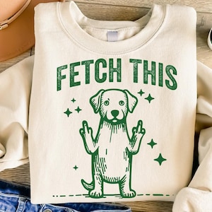 May include: A cream-colored sweatshirt with the text "FETCH THIS" in green letters. A green cartoon dog with its middle fingers raised is below the text.