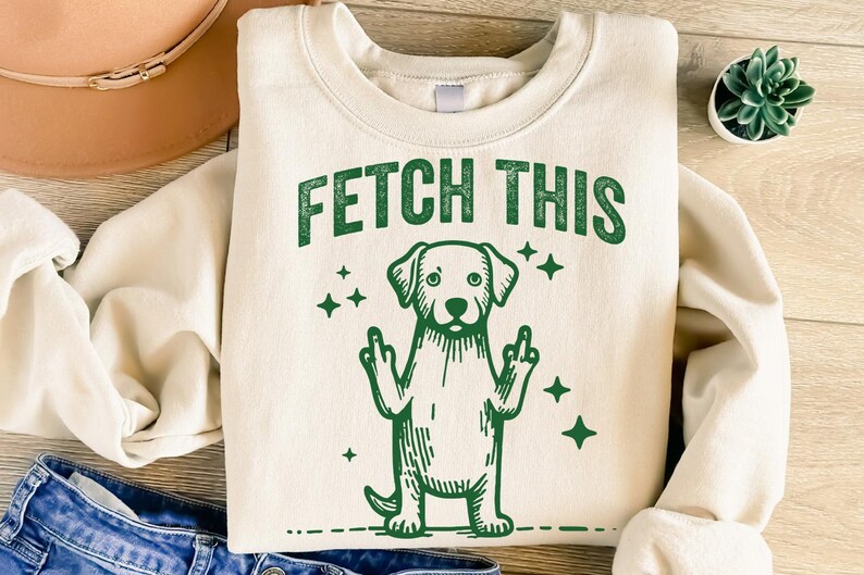 May include: A cream-colored sweatshirt with the text "FETCH THIS" in green letters. A green cartoon dog with its middle fingers raised is below the text.