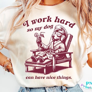 以下が含まれることがあります： 白いTシャツに、バスローブを着てサングラスをかけ、カクテルを持った犬のピンクのグラフィックが描かれています。Tシャツのテキストは「私は犬が素敵なものを手に入れられるように一生懸命働いています」です。