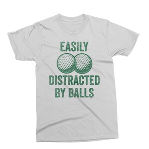 Puede incluir: Camiseta gris claro con el texto "EASILY DISTRACTED BY BALLS" en verde. Dos pelotas de golf aparecen sobre el texto. La camiseta es una prenda informal para el d&iacute;a a d&iacute;a.