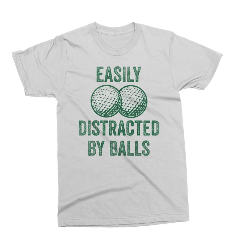 Puede incluir: Camiseta gris claro con el texto "EASILY DISTRACTED BY BALLS" en verde. Dos pelotas de golf aparecen sobre el texto. La camiseta es una prenda informal para el d&iacute;a a d&iacute;a.