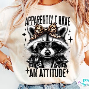 Pode incluir: Uma camiseta branca com uma ilustração em preto e branco de um guaxinim vestindo uma jaqueta de couro preta e um laço de estampa de leopardo. O texto "Aparentemente eu tenho uma atitude" está impresso na camiseta.