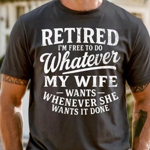 May include: Dark grey t-shirt with white text: "RETIRED I'M FREE TO DO Whatever MY WIFE WANTS - WHENEVER SHE WANTS IT DONE." The shirt has a crew neck and short sleeves.