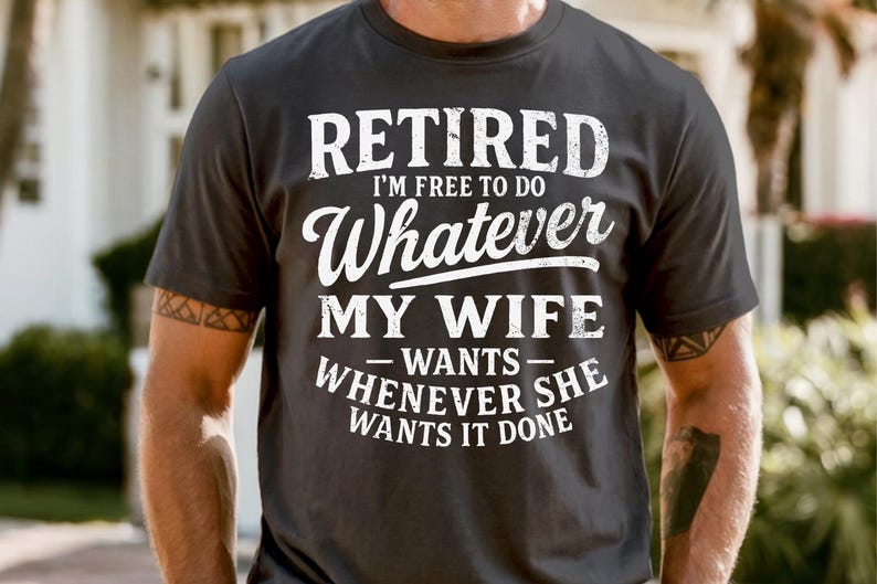 May include: Dark grey t-shirt with white text: "RETIRED I'M FREE TO DO Whatever MY WIFE WANTS - WHENEVER SHE WANTS IT DONE." The shirt has a crew neck and short sleeves.