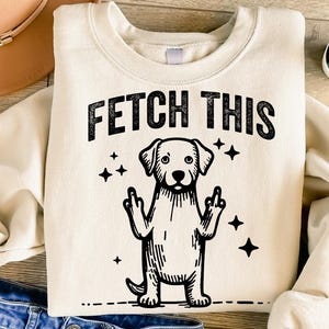 May include: A cream-colored sweatshirt with the text "FETCH THIS" in black lettering. A black and white illustration of a dog with its middle fingers up is below the text.