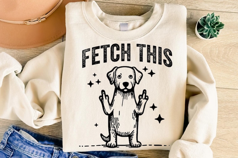 May include: A cream-colored sweatshirt with the text "FETCH THIS" in black lettering. A black and white illustration of a dog with its middle fingers up is below the text.