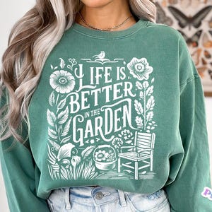May include: A green sweatshirt with a white graphic design that says "Life is better in the garden". The graphic includes flowers, leaves, a bird, and a chair.
