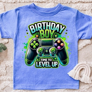 May include: A light blue t-shirt with a green, black, and white graphic of a video game controller. The text "Birthday Boy" and "Time to Level Up" is printed on the shirt.