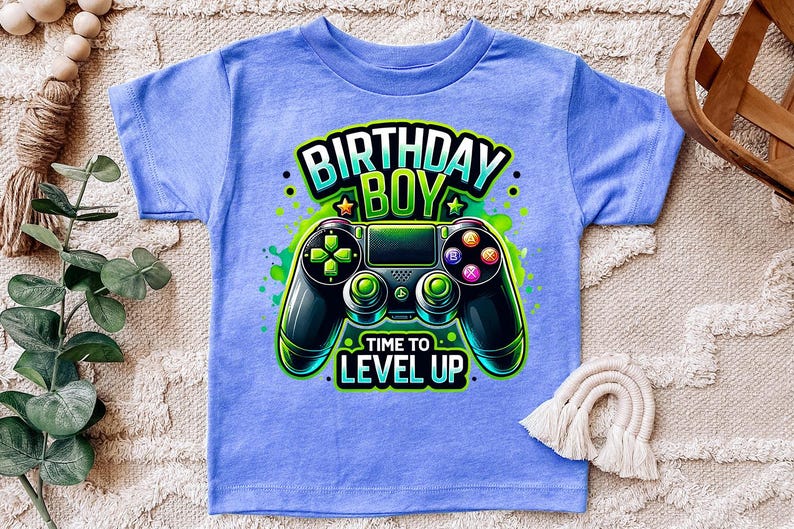 May include: A light blue t-shirt with a green, black, and white graphic of a video game controller. The text "Birthday Boy" and "Time to Level Up" is printed on the shirt.