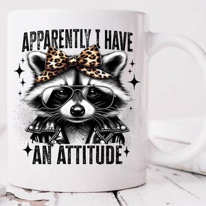 May include: White ceramic mug featuring a black and white illustration of a raccoon wearing a leather jacket and a leopard print bow. The text "Apparently I have an attitude" is printed on the mug.