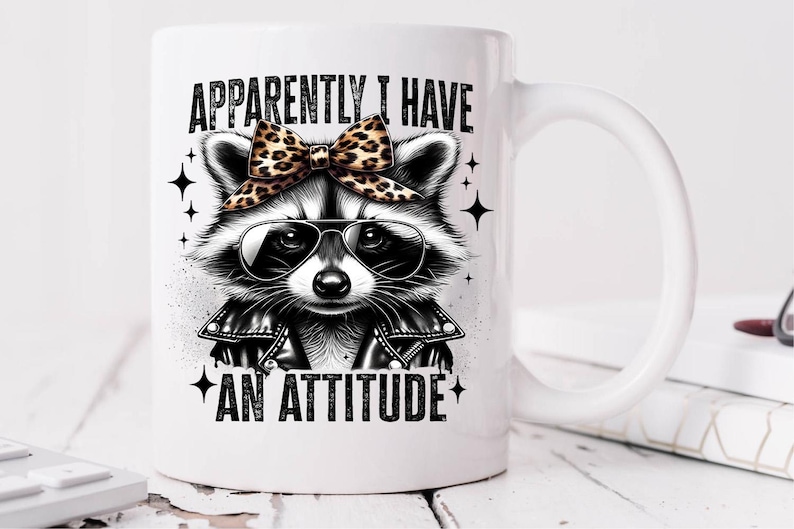 May include: White ceramic mug featuring a black and white illustration of a raccoon wearing a leather jacket and a leopard print bow. The text "Apparently I have an attitude" is printed on the mug.