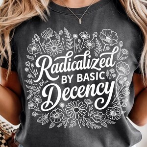 May include: Dark gray t-shirt with the text "Radicalized by Basic Decency" in white script and floral design. The shirt is a crew neck and has short sleeves. The design features various flowers and leaves.