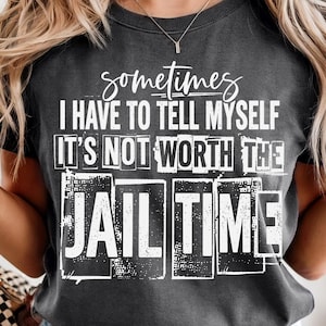 Può includere: Una t-shirt a maniche corte grigio antracite con testo bianco che recita: sometimes I HAVE TO TELL MYSELF IT'S NOT WORTH THE JAIL TIME. Il design grafico utilizza un mix di caratteri corsivi e in grassetto con un aspetto usurato e testurizzato.